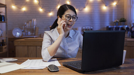 Wall Mural - business deadline and technology concept. smiling young asian chinese businesswoman with paper and laptop computer calling on smartphone at night home kitchen. friendly girl worker talking cellphone