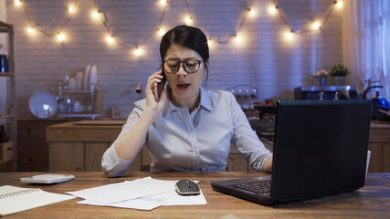 Wall Mural - Angry female manager yelling at mobile phone at dining table in late night home kitchen. girl worker disagree and argue with team partner on cellphone while working on deadline project in evening.