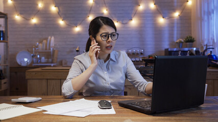 Wall Mural - Angry female freelance worker at home kitchen workplace at dark night. Serious woman frowning talking on mobile phone using laptop computer. Working hard small business and technology concept.