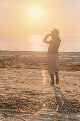 Canvas Print - grainy film effect on the picture. silhouette of a woman in a hat and dress walking along the beach against the backdrop of the setting sun