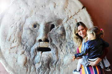 Smiling young mother with her daughter having fun at Mouth of truth  in Rome, Italy - Mum and daughter enjoy life traveling in the eternal city - Fun situation - putting the hand in the mouth of truth