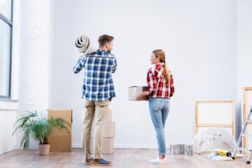 Canvas Print - back view of young couple holding cardboard box and carpet roll at home