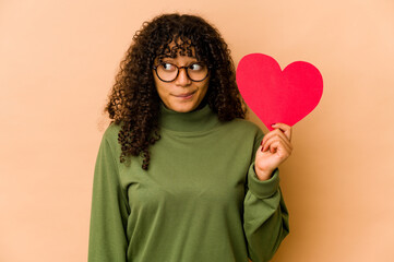 Wall Mural - Young african american afro woman holding a valentines day heart confused, feels doubtful and unsure.