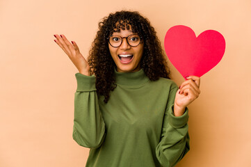 Wall Mural - Young african american afro woman holding a valentines day heart receiving a pleasant surprise, excited and raising hands.