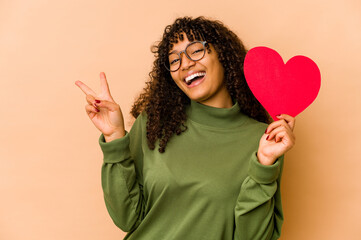 Wall Mural - Young african american afro woman holding a valentines day heart joyful and carefree showing a peace symbol with fingers.