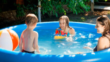 SMiling Little Boy Playing With Isster Mother With Toy Ship In Inflatable Swimming Pool At House Backyard Garden. Concept Of Happy And Cheerful Family Summer Holidays And Vacation