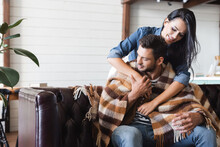 Smiling Brunette Woman Embracing Boyfriend Sitting On Leather Couch Under Plaid Blanket