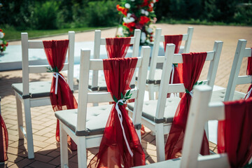 White wooden chairs decorated with red fabric and ribbons for wedding reception outdoor. Guest chairs in rows in the summer park, blurred background.