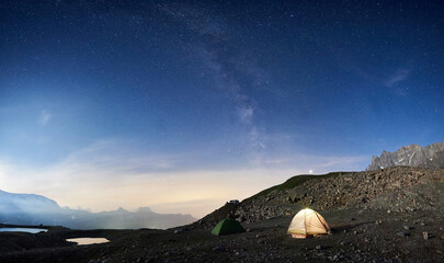 Magic scenery at night with blue sky and shining stars, beautiful mountains area with snow and lakes. Gorgeous mountain ridge with high rocky peaks, two colorful tourist tents. Panoramic view