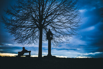 silhouette of a mysterious hacker. Silhouette portrait of a lonely anonymous man sitting on a bench as dark clouds above him roll, with the sun beginning to set behind 