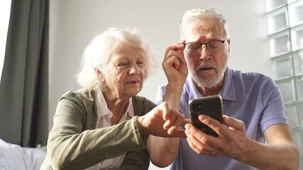 An elderly couple sits at home on a bed and understands together modern technologies, the Internet In a smartphone. users concept of the older generation.