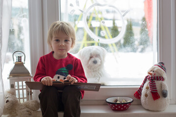 Wall Mural - Cute blond toddler child, sitting on the window, looking at the snow falling outside