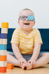 little toddler boy in lazy eye patch laughs sitting in the children's room and plays with cubes