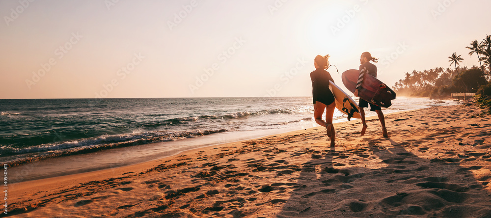 Young beautiful couple walking along the sandy beach near the ocean at sunset with surfboards, outdoor activities and sports holidays - obrazy, fototapety, plakaty 