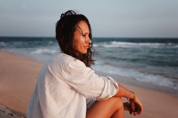 Wall Mural - Young smiling beautiful brunette woman in white shirt sitting on the beach, shot in profile