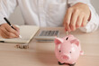 © KMPZZZ - Close up hand of young asian businessman puts a coin into the piggy bank to calculate and financial plans to spend enough money on his income for saving money.