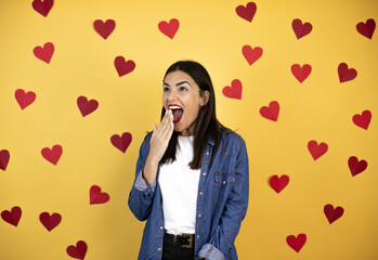 Young caucasian woman over yellow background with red hearts covering mouth with hand, shocked and afraid for mistake. surprised expression