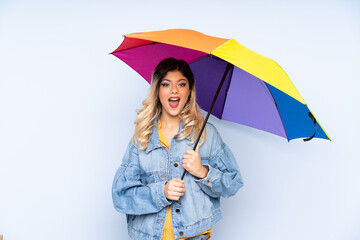 Teenager russian girl holding an umbrella isolated on blue background with surprise and shocked facial expression