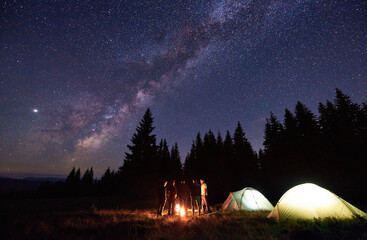 Group of five tourists are standing around burning campfire near two tents against backdrop pine forest under starry sky. Dark night sky is strewn with bright stars and Milky Way is visible on it.