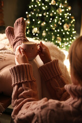 Sticker - Woman holding cup of delicious hot drink near Christmas tree indoors, closeup