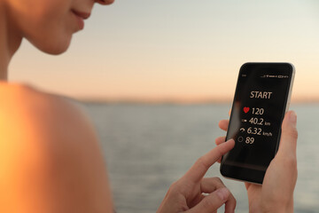 Wall Mural - Young woman using fitness app on smartphone near river at sunset, closeup