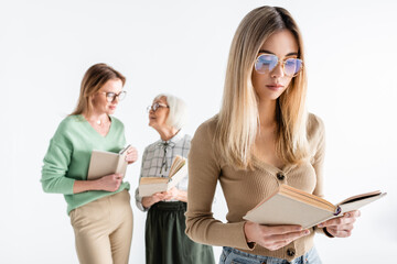 Sticker - young woman in glasses reading book near mother and granny on blurred background isolated on white