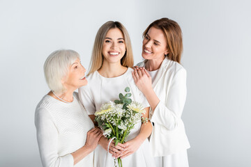 Sticker - cheerful young woman smiling while holding flowers near mother and granny isolated on grey