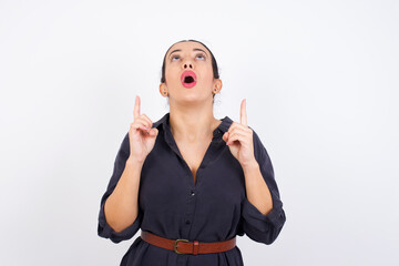 Wall Mural - young beautiful Arab woman wearing gray dress against white studio wall being amazed and surprised looking and pointing up with fingers showing something strange.