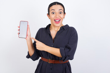 Wall Mural - Excited young beautiful Arab woman wearing gray dress against white studio wall holding and pointing with finger at smartphone with blank screen. Advertisement concept.