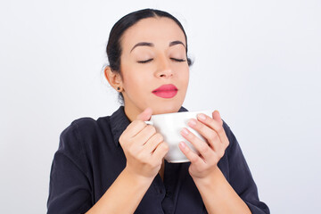 Wall Mural - young beautiful smiling Arab woman wearing gray dress against white studio background holding a cup of coffee or tea and enjoys aromatic cappuccino.
