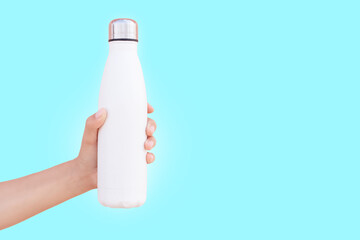 Close-up of female hand holding white reusable steel thermo water bottle isolated on background of cyan color with copy space.