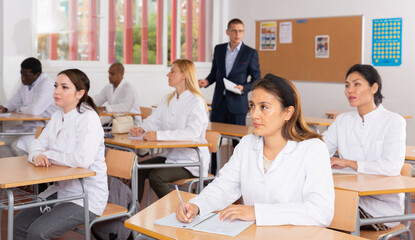 Wall Mural - Young woman in white medical coat sitting at desk in classroom attending seminar