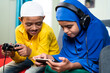 © WESTOCK - Concept of Kids busy with modern day technology lifestyle and distraction - Two muslim siblings, Brother playing game using joystick and sister playing games on mobile phone with headphones at home.