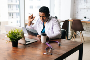 Poster - Skilled African American male doctor wearing surgeon medic suit studying history disease of patient, working laptop background of large window. Concept of medicine and health care.