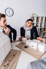 Wall Mural - happy arabian businesswoman pointing at model of green energy station near business partner, blurred foreground