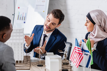 Wall Mural - translator pointing at models of building and green energy station near arabian and african american partners, blurred foreground