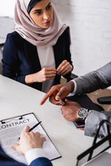 Wall Mural - arabian businesswoman looking at african american businessman pointing with finger at contract near translator, blurred foreground