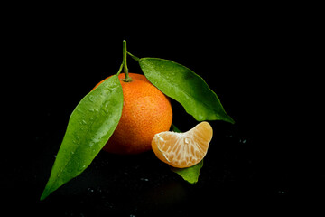 One piece of fresh and ripe tangerine with green leaves and cross section againts black background.