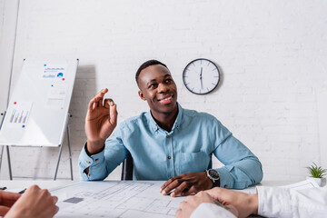 Sticker - cheerful african american businessman showing okay gesture near blueprint and business partners on foreground