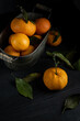 © matyuschenko - Several ripe tangerines with leaves in a metal container lie on a black table against a dark background. Rustic style with copyspace.