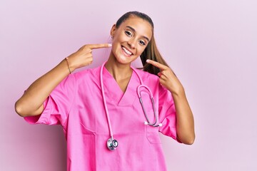 Wall Mural - Young hispanic woman wearing doctor uniform and stethoscope smiling cheerful showing and pointing with fingers teeth and mouth. dental health concept.