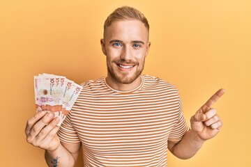 Wall Mural - Young caucasian man holding 10 colombian pesos banknotes smiling happy pointing with hand and finger to the side