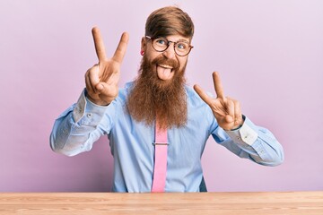 Poster - Young irish redhead man wearing business shirt and tie sitting on the table smiling with tongue out showing fingers of both hands doing victory sign. number two.