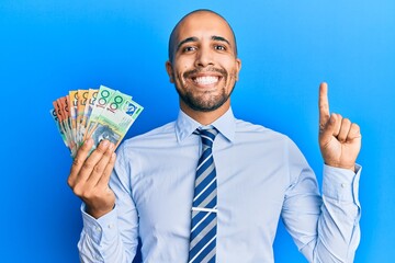 Sticker - Hispanic adult man holding australian dollars smiling with an idea or question pointing finger with happy face, number one