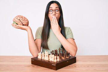 Poster - Young caucasian woman sitting at the desk playing chess holding brain covering mouth with hand, shocked and afraid for mistake. surprised expression