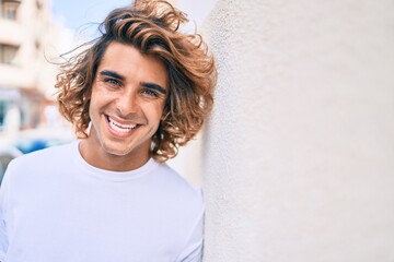 Wall Mural - Young handsome hispanic man smiling happy leaning on the wall at street of city