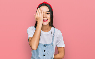 Young hispanic girl wearing casual clothes covering one eye with hand, confident smile on face and surprise emotion.