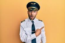 Handsome Hispanic Man Wearing Airplane Pilot Uniform Surprised Pointing With Finger To The Side, Open Mouth Amazed Expression.