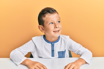 Wall Mural - Adorable caucasian kid wearing casual clothes sitting on the table looking away to side with smile on face, natural expression. laughing confident.