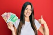 © Krakenimages.com - Young brunette woman holding norwegian krone banknotes smiling happy and positive, thumb up doing excellent and approval sign